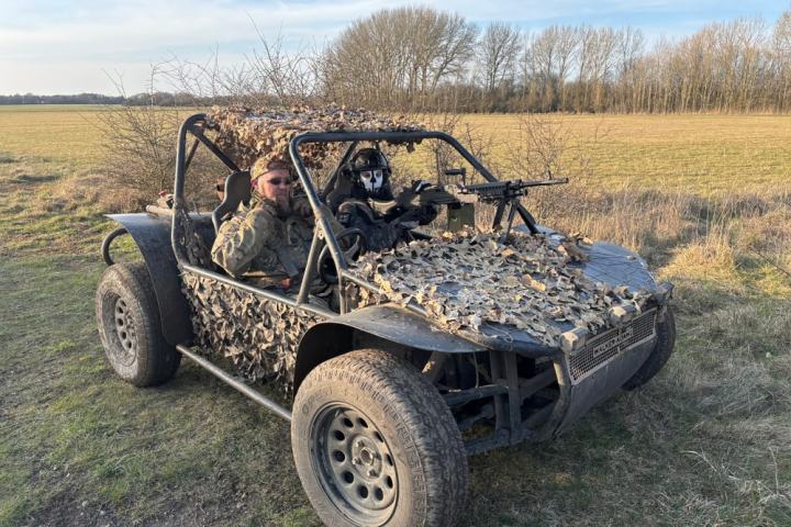 Two people in camo-covered buggy with mounted rifle in an open field.