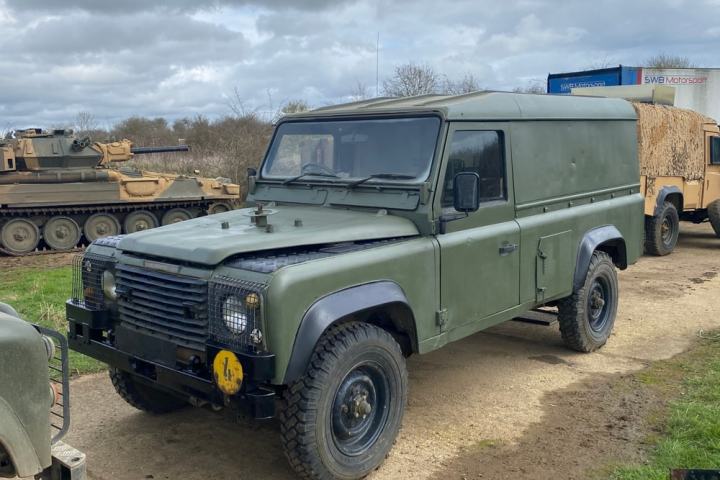 Green military vehicle parked beside a tank on a dirt road, under cloudy skies.